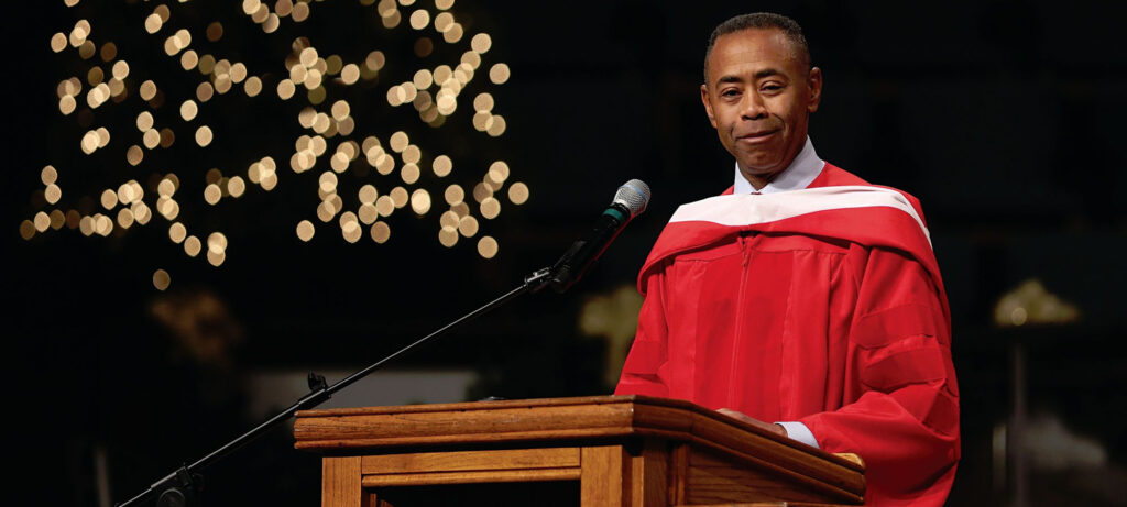 A man in a red academic gown, possibly Reggie Thomas, stands at a podium with a microphone, appearing to speak. Out-of-focus lights shine in the background, suggesting a sense of union or family gathering.