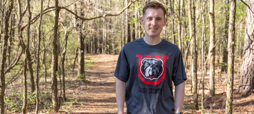 A person stands on a wooded path, wearing a dark gray Dogs Country T-shirt with a bulldog graphic and red U. Sunlight filters through the trees as they channel Tristan Kelly’s spirit, running toward redemption.
