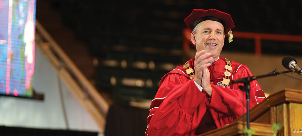 A person in academic regalia, including a red gown and cap with a gold medallion, stands at a podium, smiling and clapping during a graduation ceremony celebrating 5 years of Courage and Conviction under the Oliver administration.