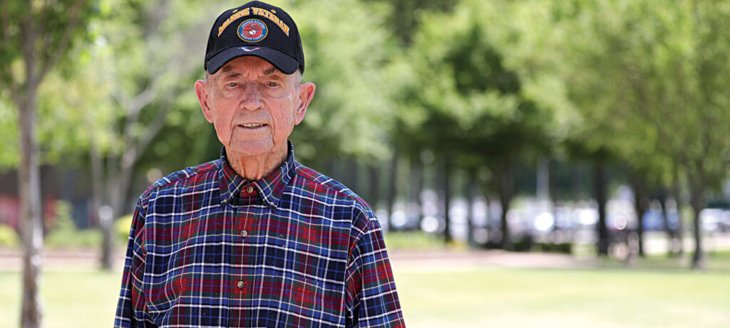 An elderly man, Roe Neal, wearing a black baseball cap with military insignia and a plaid shirt, stands outdoors in a park, surrounded by trees and green grass—a moment in his life’s adventure with God.