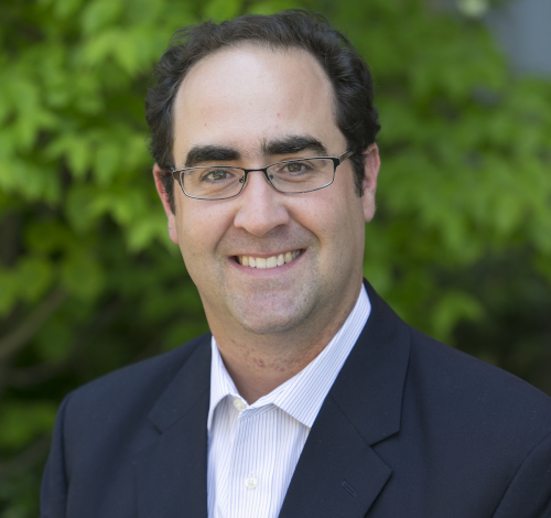 A man with short dark hair and glasses is wearing a dark suit jacket and light-colored shirt. He is smiling and standing outdoors in front of green foliage, perhaps at the Spring Bible Conference where Gignilliat spoke on Ecclesiastes.