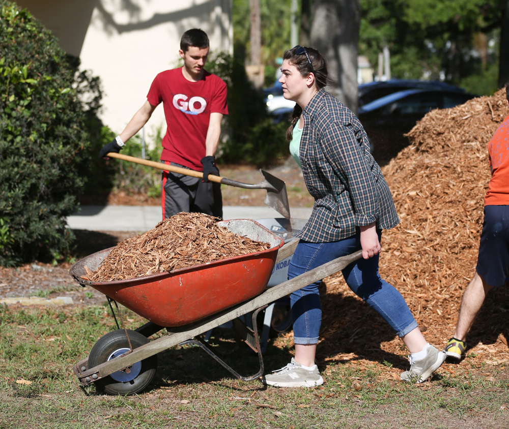 Union students help push a wheelbarrow filled with wood chips or mulch while another person shovels near a large mulch pile outdoors on a sunny day, supporting the Tampa church plant project.
