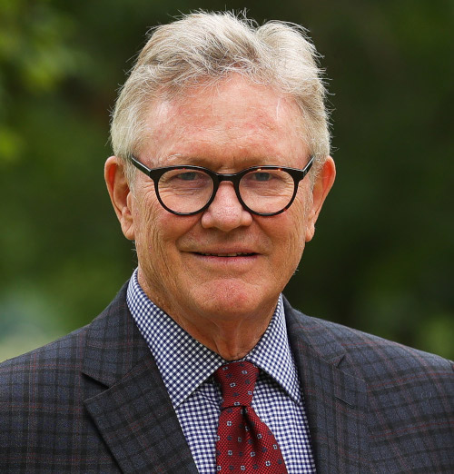 An older man with gray hair and glasses is wearing a dark checkered suit, patterned shirt, and red tie, standing outdoors after the Union summer commencement, with greenery in the blurred background.