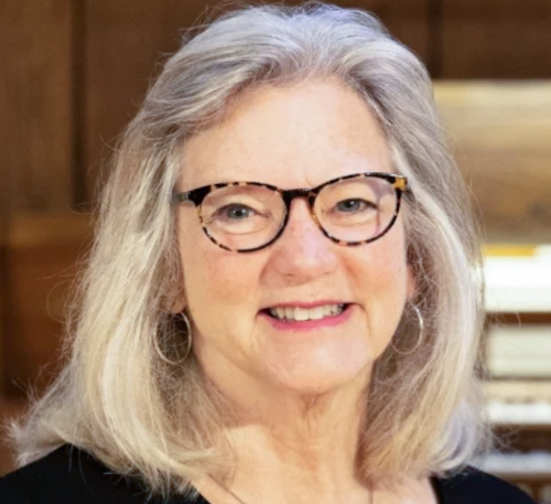 An older woman with gray hair, wearing glasses and hoop earrings, smiles at the camera. She is indoors in front of a wooden wall, with books or papers possibly related to the Handford annual concert visible in the background.