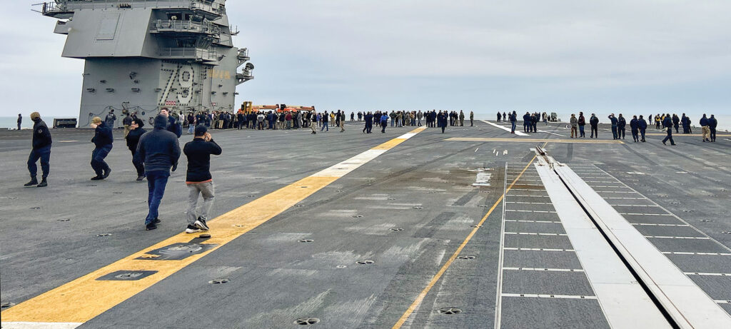 A large group of people stand and walk on the deck of an aircraft carrier under a cloudy sky, as the ship's control tower looms on the left—ready to go full speed ahead, captured in this moment by A.J. Zeak.