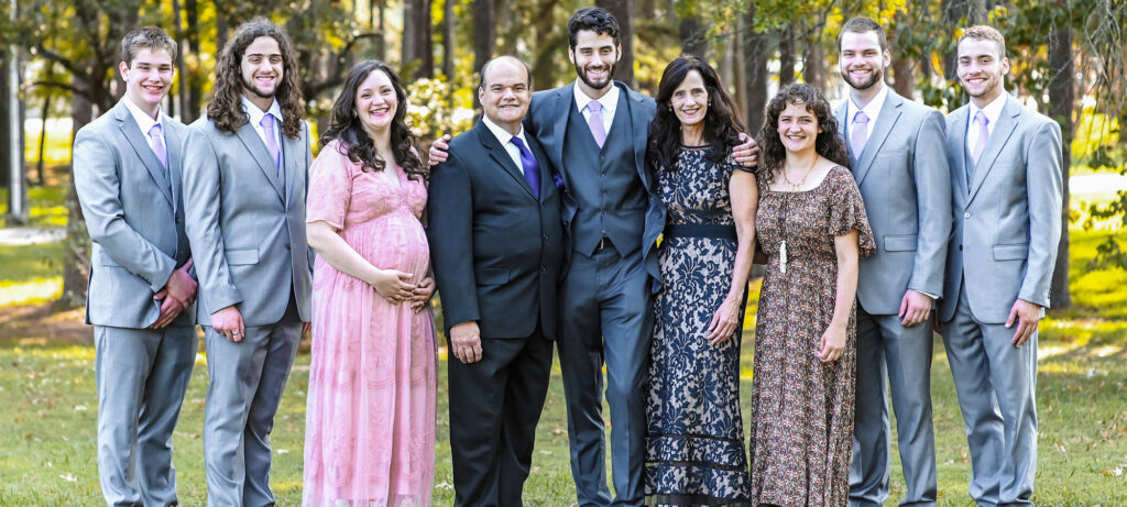 A group of nine people, including six men in gray suits, two women in dresses (one in pink and one in brown), and one man in a black suit, stand outdoors in a park, smiling at the camera—celebrating the Carbonells' family legacy and union.