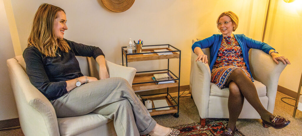 Chelsy Crawford and a friend sit facing each other in armchairs, engaged in conversation in a well-lit room. A cart with office supplies is visible between them, making space feel inviting and both appear relaxed.