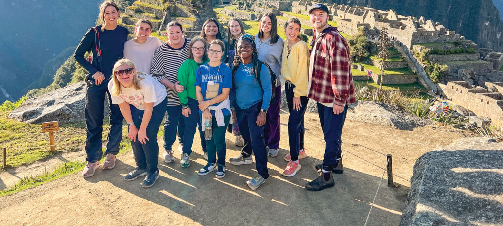 A group of thirteen people from the EDGE Program stand and smile for a photo at Machu Picchu, with ancient stone ruins and green mountains visible in the background under bright sunlight.