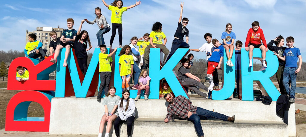 A group of children and teens from a micro school pose on and around large colorful letters spelling “DR MLK JR” outdoors on a sunny day, celebrating educational independence. Buildings and trees are visible in the background.