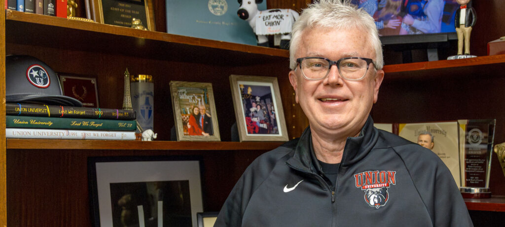 A man with short gray hair and glasses, identified as Robbie Graves, stands in front of a wooden bookshelf filled with framed photos, awards, and memorabilia. He is wearing a black Union University jacket and smiling with encouragement at the camera.
