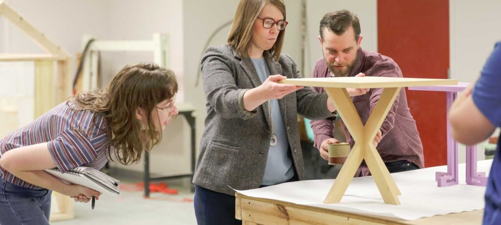 Three people examine and adjust a small wooden table on a workbench. One person crouches and takes notes while the other two hold and inspect the table. Tools and materials are visible in the background.