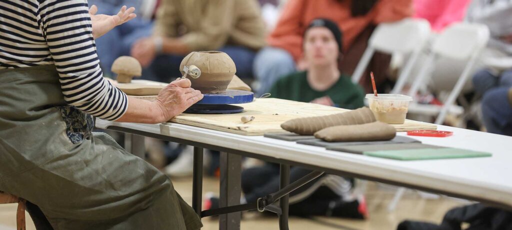 A person wearing a striped shirt and apron demonstrates pottery techniques at a table with clay, tools, and a bowl, while seated people watch in the background.