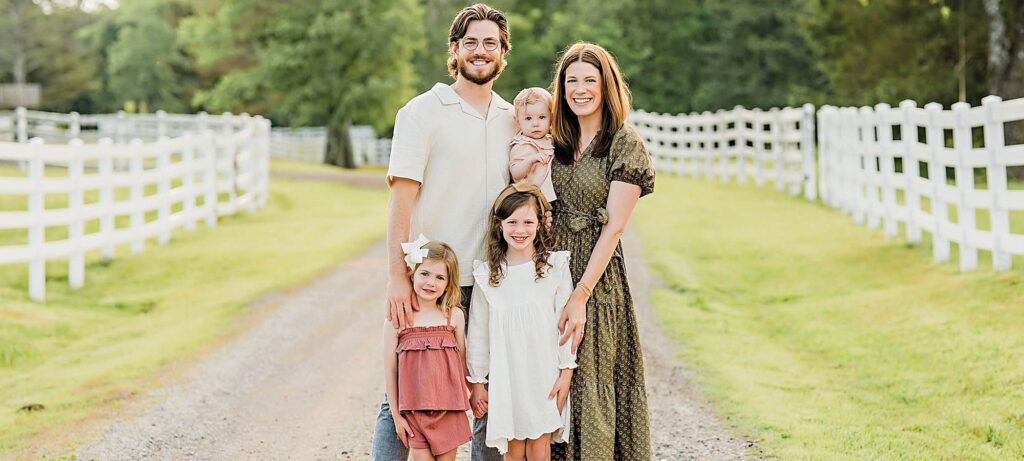Clayton and McKinley Martin stand on a dirt path between white fences, trees in the background. The parents hold a baby, while two young girls stand in front, all smiling at the camera—capturing a moment Beyond the Degree.