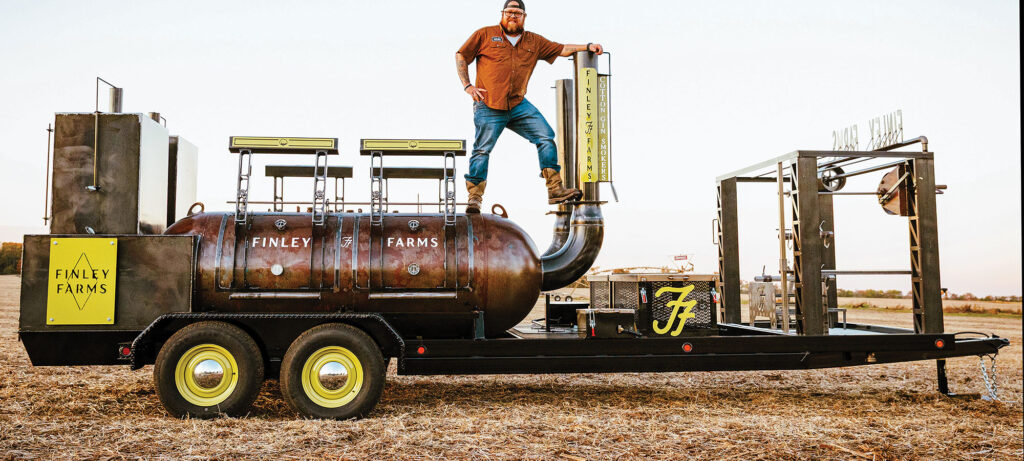 A man stands on a large, custom barbecue smoker mounted on a trailer in a field. The smoker, labeled “Finley Farms,” features three smoking compartments, a tall temperature gauge, and craftsmanship refined by fire.