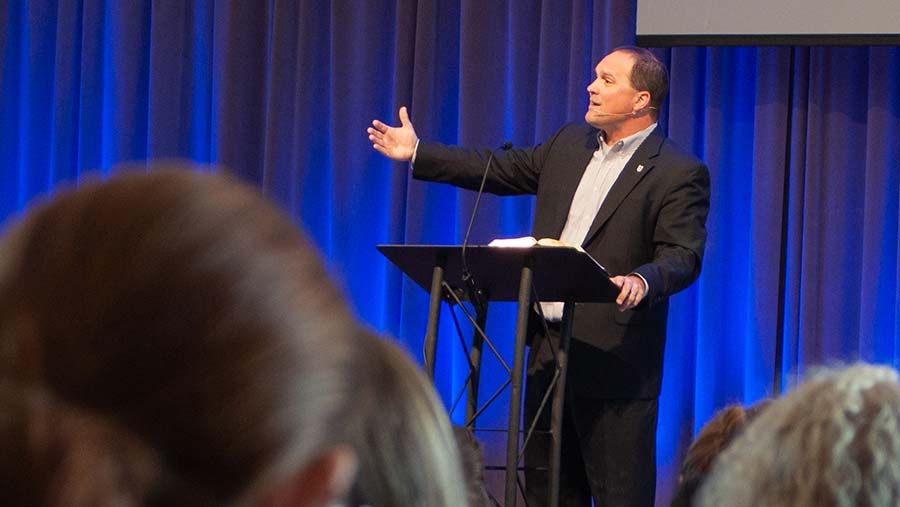 A man in a suit stands at a podium, speaking and gesturing with one hand extended, offering sermon tips to the audience before him. Blue curtains provide a backdrop as listeners in the foreground engage with his advice.