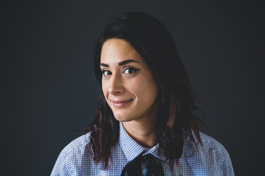 Elena Rodríguez, with medium-length dark hair, wearing a collared shirt and a black ribbon tie, looks at the camera with a slight smile against a plain dark background.