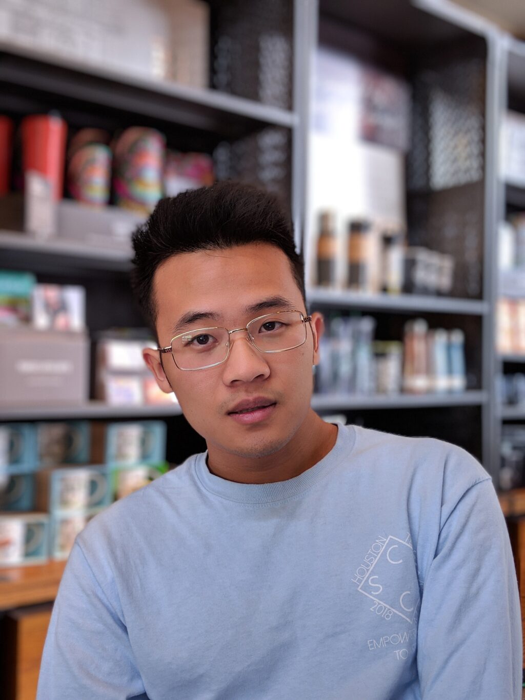 A young man with short black hair and glasses wearing a light blue shirt is sitting indoors in front of shelves stocked with various colorful products.