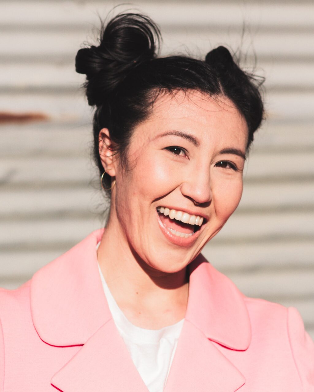 Isla Williams, a woman with dark hair in two buns, is wearing a pink jacket and smiling widely. The background is out of focus with horizontal lines.