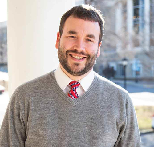 A man with short brown hair and a beard is standing outside, wearing a grey sweater over a white dress shirt with a red striped tie, smiling at the camera at the Union’s Creative Writing Workshop.