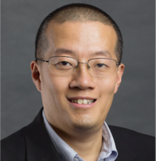 A man with short hair and glasses, wearing a dark suit jacket and light-colored collared shirt, smiles at the camera in front of a plain gray background—perhaps preparing for the Powell Missions Lecture at IMB with Hou.