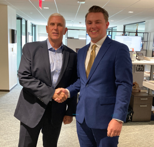 Two men in business suits shake hands and face the camera while standing in an office, possibly discussing a summer internship, with desks, computers, and overhead lights visible in the background.