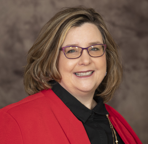 A woman with shoulder-length light brown hair wearing glasses, a red blazer, and a black top smiles in front of a brown, blurred background, representing the College of Nursing at Health Sciences.