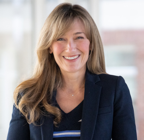 Janney, a woman with long, light brown hair, smiles at the camera in a navy blazer over a striped top. She stands in a bright indoor setting with blurred windows, inviting you to the Carls-Schwerdfeger History Lectures on Oct. 6.