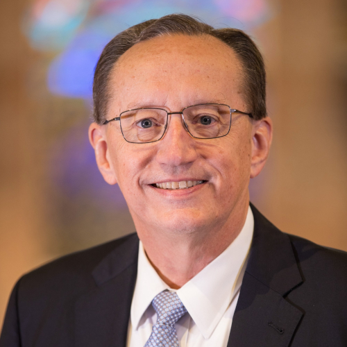 A middle-aged man with short brown hair and glasses is smiling at the camera, dressed for an SBC Union graduation. He wears a dark suit jacket, white dress shirt, and light blue tie. The background is softly blurred with colorful lights.
