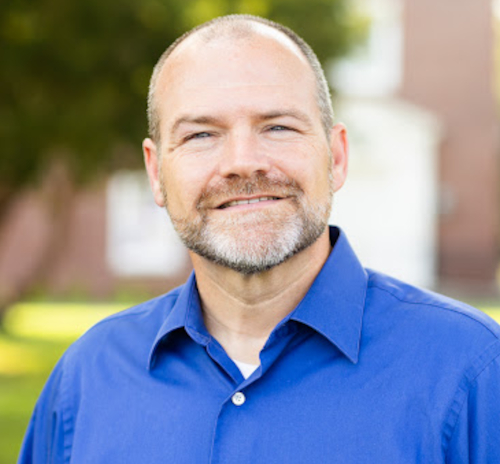 A middle-aged man with a short beard and a shaved head is wearing a blue collared shirt, standing outdoors with greenery and a building in the blurred background at the Jan. 30 W.D. Powell Missions Lecture featuring Bledsoe.