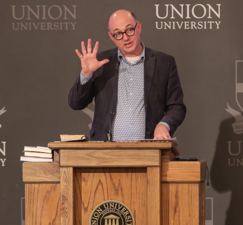 A man wearing glasses and a blazer stands at a wooden podium with the Union University logo, gesturing with his left hand raised while speaking about the Beatitudes during the Spring Bible Conference. Several books and a notebook are on the podium.