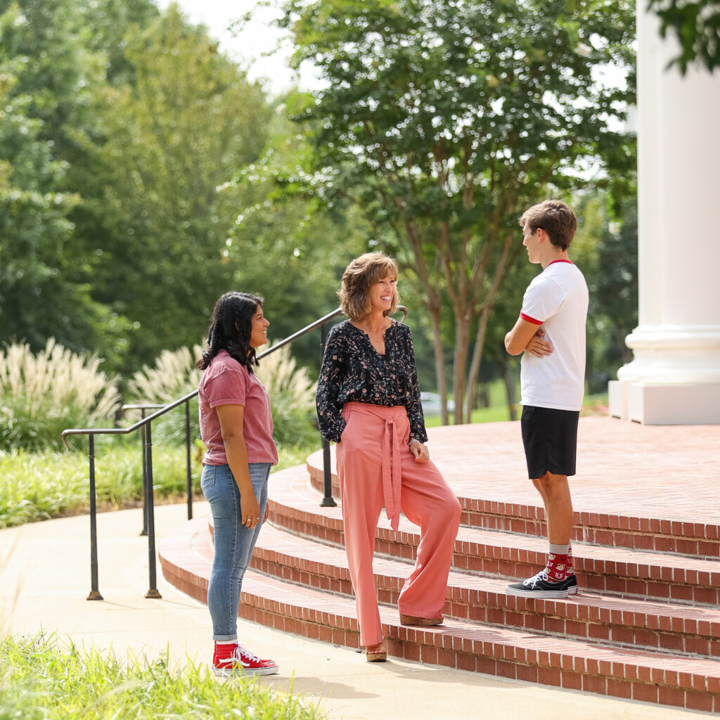 Three people stand and talk near brick steps outside, possibly discussing trustees or budget matters. A woman in pink pants and a floral blouse faces a young woman in jeans and a young man in a white shirt, with green trees and grass in the background.