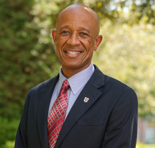 A man in a dark suit, light blue shirt, and red patterned tie stands outdoors, smiling at the camera. The background is blurred greenery and trees with sunlight filtering through. Lewis, MCUTS dean at Union University, radiates warmth and professionalism.