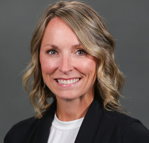 A woman with wavy blonde hair, Dean Jones of the College of Pharmacy, smiles at the camera in a black blazer over a white top against a plain gray background.