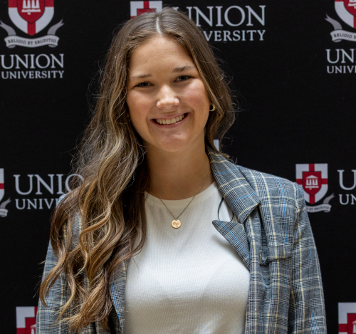 A woman with long, wavy brown hair stands in front of a Union University backdrop, smiling. Representing Gibson County, she wears a plaid blazer over a white top and round gold necklace—celebrating her Union academic scholarship.