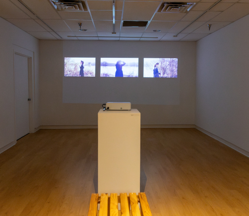 A small gallery room with white walls and a wooden floor features a projector on a pedestal, displaying four video stills of a person outdoors on the far wall. Part of the Lee exhibit, this installation will be on display through Jan. 30. A wooden bench is in the foreground.