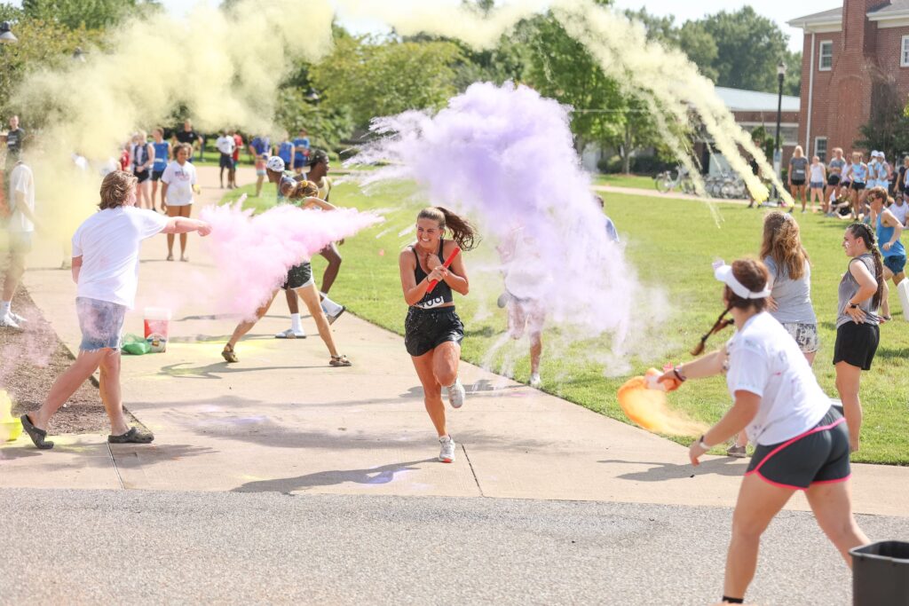 A group of people outdoors participate in a color run event, throwing clouds of yellow, purple, and pink powder as a woman in athletic clothing runs through the colorful scene.