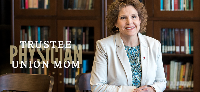 A woman with curly hair in a light blazer and patterned blouse stands in front of bookshelves. Text on the image reads: Trustee, Physician, Union Mom. This smiling physician rests her hands on the back of a chair.