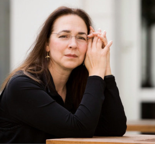 A person with long brown hair and glasses sits at a wooden table, resting their chin on their hands, looking at the camera with a neutral expression. They are outdoors at Union Lyceum, wearing a black top.