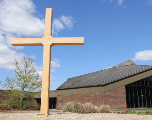 A large yellow cross stands outside a modern brick and metal building in Memphis, likely a church offering a Master of Arts in Bible and Theology, with blue sky above. Shrubs and a small tree are near the entrance.
