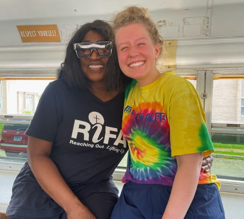Two women are smiling and posing together inside the Bus Stop Café bus. One, a Union PR major, wears dark sunglasses and a black RIFA t-shirt; the other sports a yellow tie-dye t-shirt. A sign above them reads Respect Yourself.
