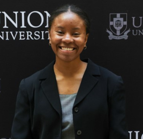 A person wearing a black blazer and gray top is smiling in front of a black backdrop with partial text and logos for Union University, celebrating their Madison Union academic scholarship.