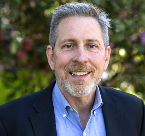 A middle-aged man with gray hair and a beard, dressed in a blue shirt and dark blazer, smiles at the camera. He is standing outdoors amid greenery and flowers, possibly attending the Union’s Constitution Day lectures.