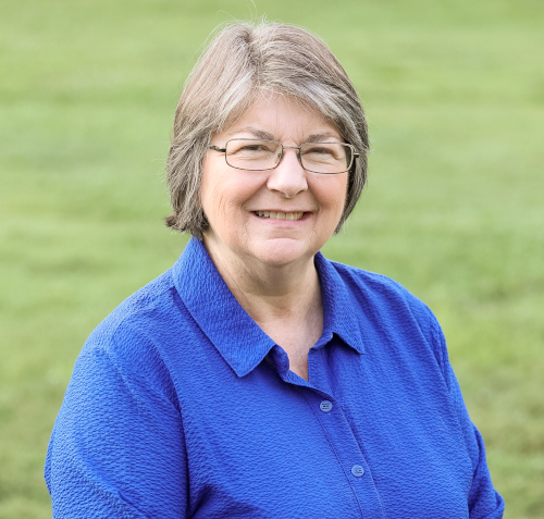 An older woman with gray hair and glasses is wearing a bright blue shirt and smiling at the camera. She is outdoors on a grassy lawn, possibly attending a Union summer commencement or listening to a commencement address.
