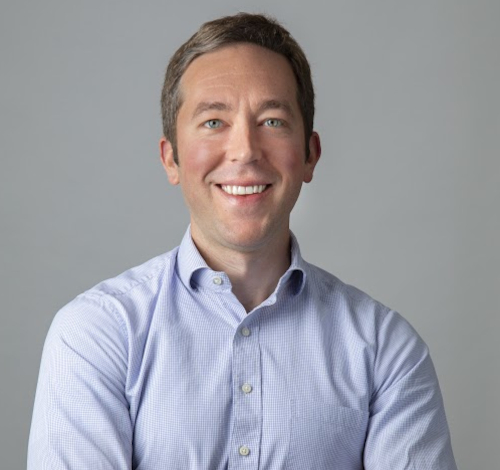 A man with short brown hair, wearing a light blue checkered button-up shirt, stands with his arms crossed and smiles at the camera against a plain gray background, ready to attend the Union luncheon event for Faith in the Marketplace.