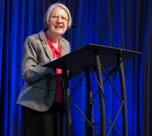 An older woman wearing glasses, a gray blazer, and a pink blouse stands at a podium with a microphone, speaking about the Union. A dark blue curtain hangs in the background as she accepts the Dodd Award.