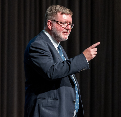 A man in a suit and tie is standing and pointing with his right hand. With short gray hair, glasses, and a beard, he resembles a Yale historian discussing British imperialism. The background is dark with vertical curtains.