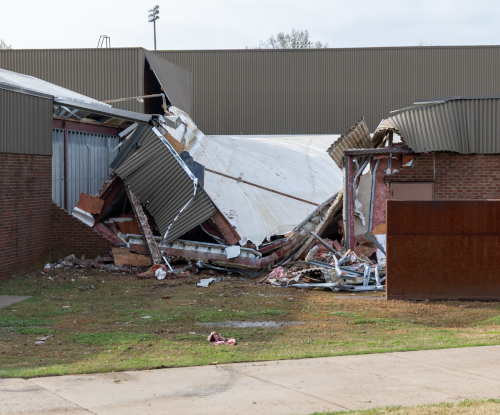 A Union building with a collapsed metal roof and damaged walls shows storm damage, with debris scattered outside. The structure appears partially destroyed, exposing beams and insulation, leading to a partial closure.