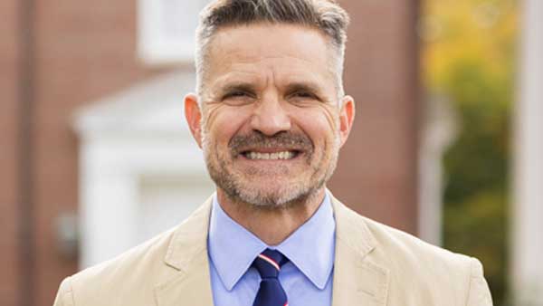 A middle-aged man with short gray hair and a trimmed beard is smiling. He is wearing a light tan blazer, a blue dress shirt, and a blue and pink striped tie—ready to attend the upcoming 2027 Bible Conference. A blurred building is in the background.