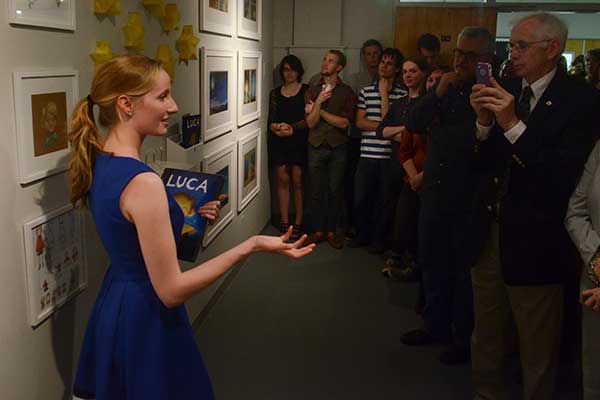 A woman in a blue dress stands near framed artwork on a wall, holding a book and speaking to a group of people. One person in the crowd takes a photo with a phone. Yellow star-shaped decorations are visible in the background.
