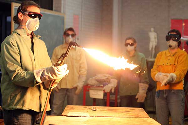 A person wearing protective gear uses a torch that emits a flame, while three others in safety goggles and masks observe in a workshop setting.